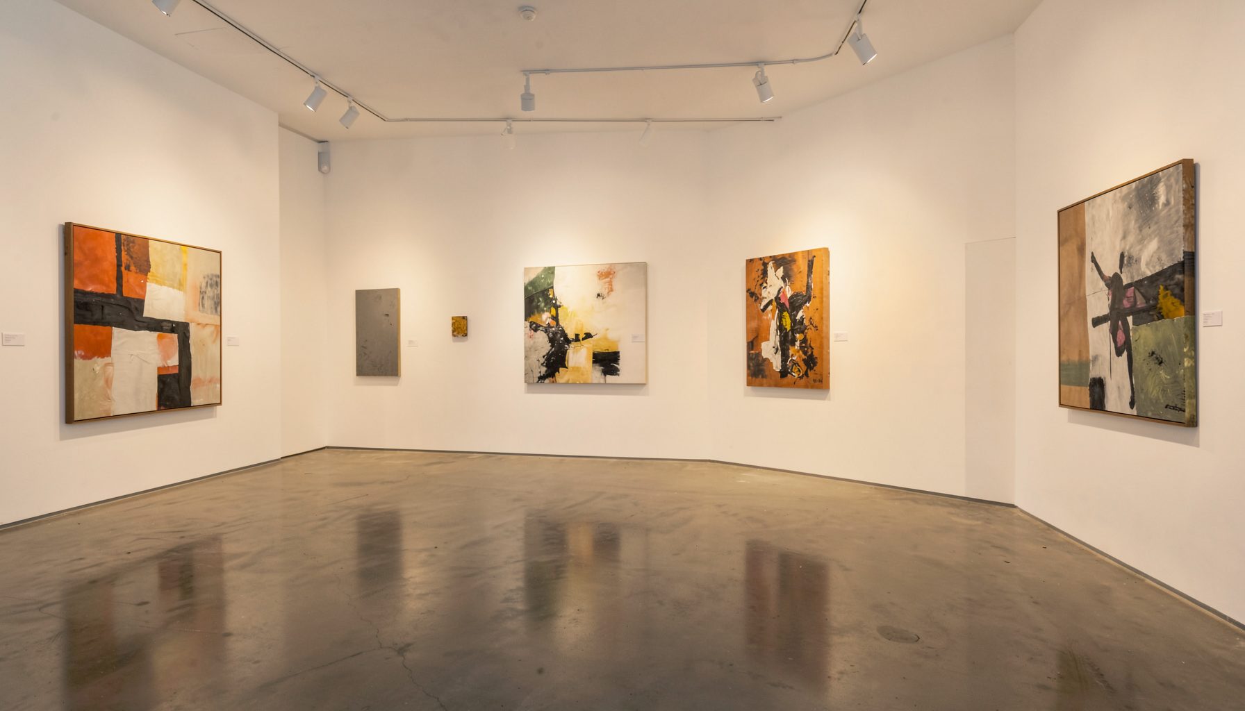 A warmly lit contemporary gallery interior with white walls, polished concrete floors, and abstract paintings spaced along the walls