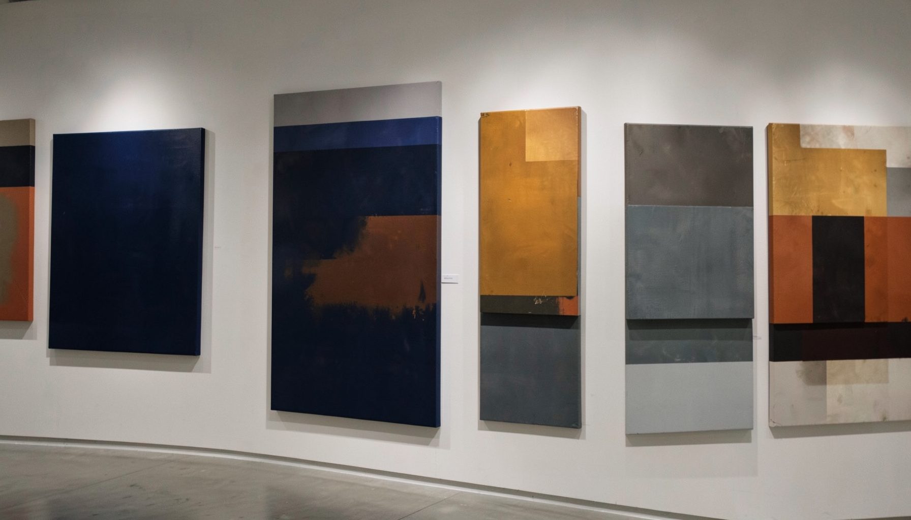 An abstract gallery wall with layered canvases in deep navy, amber, and slate tones, softly lit from above